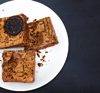 Three square brownies are placed on a white plate against a dark background. One of the brownies is topped with a sandwich cookie, while the other two have visible chocolate chips and a cracked surface.