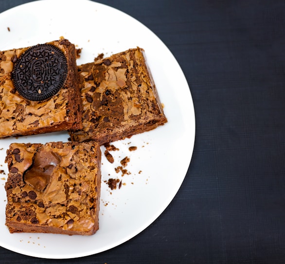 Three square brownies are placed on a white plate against a dark background. One of the brownies is topped with a sandwich cookie, while the other two have visible chocolate chips and a cracked surface.