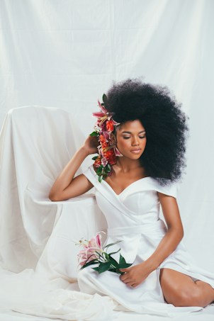 A woman in a white dress with an off-shoulder design sits gracefully against a draped white background. Her hair is styled voluminously and adorned with vibrant red and pink flowers, matching the bouquet of lilies she holds in her hand. Her eyes are closed, and her expression is serene, exuding elegance and calmness.