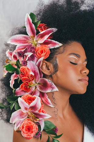 A woman with closed eyes is adorned with a vibrant floral arrangement intertwined in her voluminous, curly hair. The flowers include pink lilies, red roses, and green leaves, creating a harmonious and colorful contrast. Her makeup is minimal, with white eyeliner accentuating the serene expression on her face.
