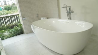 A modern bathroom features a large, freestanding white bathtub with a sleek faucet. The bathroom is partially open to the outside, providing a view of greenery and letting natural light flood in. There is a tiled wall with a mounted toilet and subtle beige tiles on the floor.