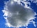 A large, fluffy cloud occupies most of the image, surrounded by a clear blue sky. The cloud appears dense and has a mixture of white and gray tones, with the sun illuminating the edges. Wispy smaller clouds are scattered around.