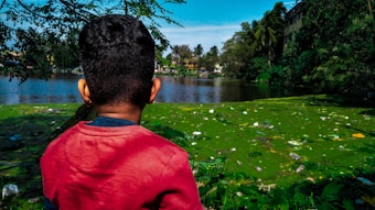 A child wearing a red shirt is facing a polluted body of water filled with algae and garbage. Surrounding the water, there are trees and palm trees with a clear blue sky in the background.