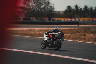 A motorcyclist wearing protective gear is riding a white sports bike on a race track. The track is surrounded by dry grass and trees, with a fence and some vehicles in the background. The rider appears to be leaning into a turn.