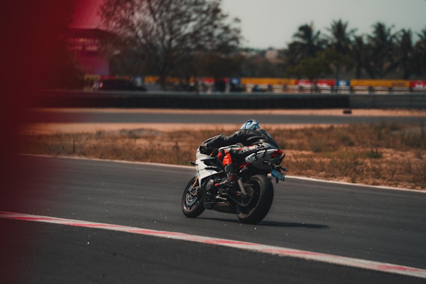A motorcyclist wearing protective gear is riding a white sports bike on a race track. The track is surrounded by dry grass and trees, with a fence and some vehicles in the background. The rider appears to be leaning into a turn.