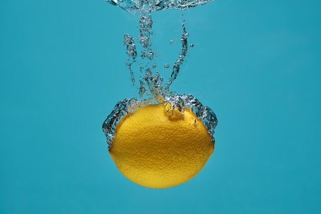 A lemon is submerged in water with bubbles forming around it, set against a bright blue background.