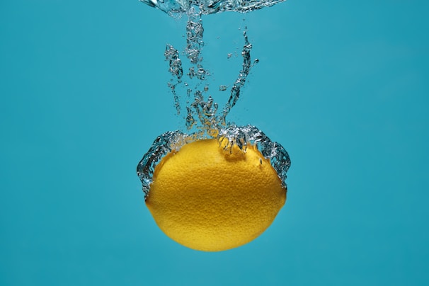 A lemon is submerged in water with bubbles forming around it, set against a bright blue background.