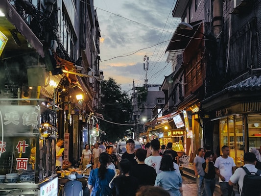 A bustling street market during evening hours, lined with diverse food stalls and shops with bright signs. People walk along the narrow street, engaging with vendors. The sky is partially cloudy, and electric wires crisscross overhead.