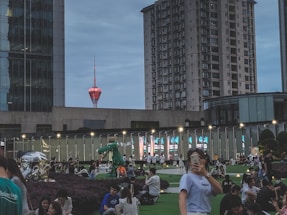 In a bustling urban scene, people are gathered in an outdoor public space with modern high-rise buildings in the background. The sky has a twilight hue, and a distinctive red tower stands prominently in the distance. Many individuals are sitting on green lawns, socializing or using their phones, creating a lively and communal atmosphere.