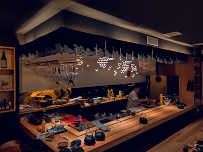 A cozy and dimly lit sushi bar features a wooden counter with various utensils and ingredients. A person is preparing food behind the counter. Glasses hang upside down from a rack above, and decorative patterns adorn the wall, adding to the ambiance.
