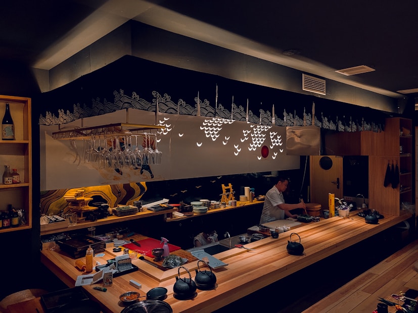 A cozy and dimly lit sushi bar features a wooden counter with various utensils and ingredients. A person is preparing food behind the counter. Glasses hang upside down from a rack above, and decorative patterns adorn the wall, adding to the ambiance.