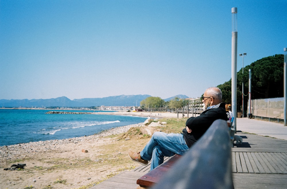 man in black jacket sitting on brown wooden bench near body of water during daytime