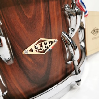 A close-up of a wooden drum with a smooth, polished finish. The drum features chrome hardware and a decorative metal badge with engraved designs. A ribbon with red, white, and blue stripes is attached to the hardware. There's a booklet or tag resting against the drum, adding a touch of elegance.