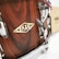 A close-up of a wooden drum with a smooth, polished finish. The drum features chrome hardware and a decorative metal badge with engraved designs. A ribbon with red, white, and blue stripes is attached to the hardware. There's a booklet or tag resting against the drum, adding a touch of elegance.