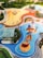 A vibrant playground with various play areas including a sandy zone with slides, a blue water-themed area with a lighthouse and boat structure, and other equipment. The playground is surrounded by green lawns, trees, and walking paths. There is also a glass-roofed structure near the edges.