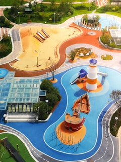 A vibrant playground with various play areas including a sandy zone with slides, a blue water-themed area with a lighthouse and boat structure, and other equipment. The playground is surrounded by green lawns, trees, and walking paths. There is also a glass-roofed structure near the edges.