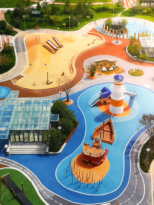 A vibrant playground with various play areas including a sandy zone with slides, a blue water-themed area with a lighthouse and boat structure, and other equipment. The playground is surrounded by green lawns, trees, and walking paths. There is also a glass-roofed structure near the edges.