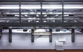 A view of a printing press with stacks of paper beneath it. The machinery is visible with metallic elements and a strong light source at the top. The scene conveys an industrial setting with a focus on mass production.