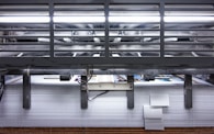 A view of a printing press with stacks of paper beneath it. The machinery is visible with metallic elements and a strong light source at the top. The scene conveys an industrial setting with a focus on mass production.