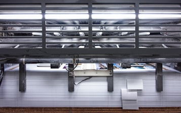 A view of a printing press with stacks of paper beneath it. The machinery is visible with metallic elements and a strong light source at the top. The scene conveys an industrial setting with a focus on mass production.