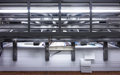 A view of a printing press with stacks of paper beneath it. The machinery is visible with metallic elements and a strong light source at the top. The scene conveys an industrial setting with a focus on mass production.