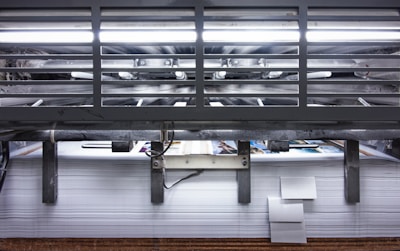 A view of a printing press with stacks of paper beneath it. The machinery is visible with metallic elements and a strong light source at the top. The scene conveys an industrial setting with a focus on mass production.