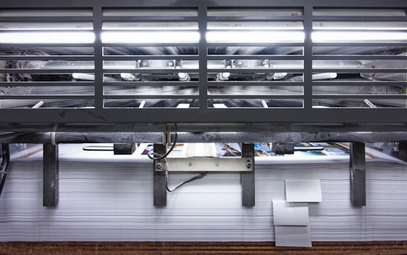A view of a printing press with stacks of paper beneath it. The machinery is visible with metallic elements and a strong light source at the top. The scene conveys an industrial setting with a focus on mass production.