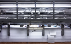 A view of a printing press with stacks of paper beneath it. The machinery is visible with metallic elements and a strong light source at the top. The scene conveys an industrial setting with a focus on mass production.