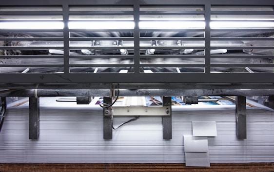 A view of a printing press with stacks of paper beneath it. The machinery is visible with metallic elements and a strong light source at the top. The scene conveys an industrial setting with a focus on mass production.