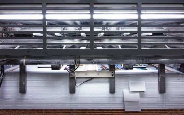 A view of a printing press with stacks of paper beneath it. The machinery is visible with metallic elements and a strong light source at the top. The scene conveys an industrial setting with a focus on mass production.