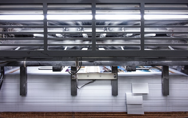 A view of a printing press with stacks of paper beneath it. The machinery is visible with metallic elements and a strong light source at the top. The scene conveys an industrial setting with a focus on mass production.