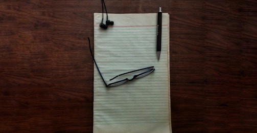 A wooden desk surface holds a yellow legal pad, upon which rests a pair of black-framed glasses, black earbuds, and a black pen.