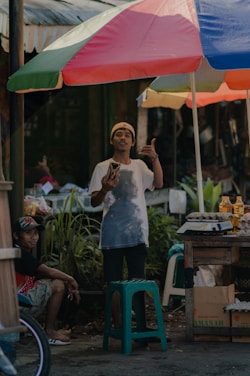 A street vendor stands under a colorful umbrella, holding a phone and gesturing with his hand. He is wearing a white T-shirt and a backwards cap. Nearby, another man sits casually on a stool, wearing a baseball cap and shorts. The stall has items such as eggs and bottles on display, with green plants and boxes visible in the background.