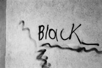 Graffiti art featuring the word 'Block' spray-painted in black on a textured concrete wall. The writing is slightly stylized, with a few extra lines and curves around it.