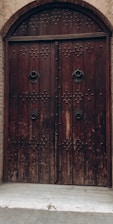 A large, arched wooden door with intricate metal studs and ornate metal handles, set into a brick wall. The door appears weathered, adding a vintage or historical feel to the architecture.