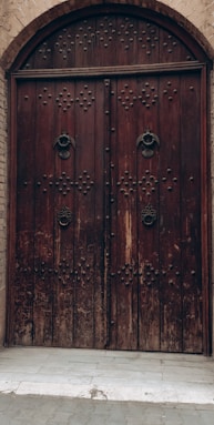 A large, arched wooden door with intricate metal studs and ornate metal handles, set into a brick wall. The door appears weathered, adding a vintage or historical feel to the architecture.