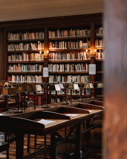 A cozy library setting featuring rows of wooden desks with reading lamps and shelves filled with books. Warm light illuminates the space, creating a comfortable and inviting atmosphere.