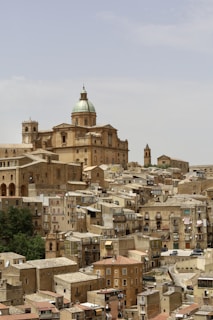 A historic townscape with densely packed, aged buildings made of stone and brick, featuring a prominent dome and bell tower in the background. The architecture showcases a Mediterranean style, typical of Southern European towns, with narrow streets and windows adorned with shutters. A few scattered trees provide greenery amidst the earth-toned structures.