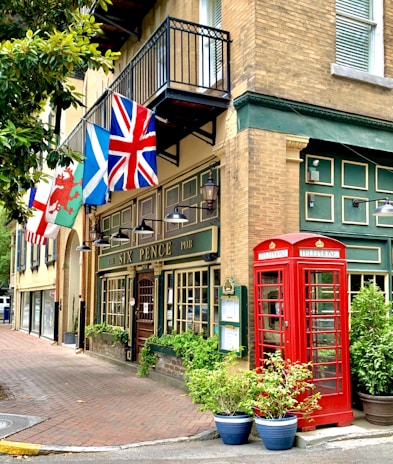 A traditional British-style pub with green accents, featuring a red telephone booth and various national flags displayed above the entrance. The facade is adorned with decorative lighting and lush green plants in pots. Large windows and a wooden door complete the charming appearance.