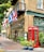A traditional British-style pub with green accents, featuring a red telephone booth and various national flags displayed above the entrance. The facade is adorned with decorative lighting and lush green plants in pots. Large windows and a wooden door complete the charming appearance.