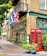 A traditional British-style pub with green accents, featuring a red telephone booth and various national flags displayed above the entrance. The facade is adorned with decorative lighting and lush green plants in pots. Large windows and a wooden door complete the charming appearance.