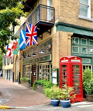 A traditional British-style pub with green accents, featuring a red telephone booth and various national flags displayed above the entrance. The facade is adorned with decorative lighting and lush green plants in pots. Large windows and a wooden door complete the charming appearance.