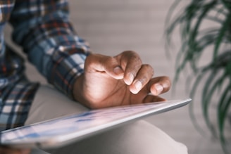A person engages with a tablet device using their fingertips. The individual is dressed in a plaid shirt, and the focus is on their hand interacting with the touchscreen. A blurred background, featuring an out-of-focus plant, provides a soft context.