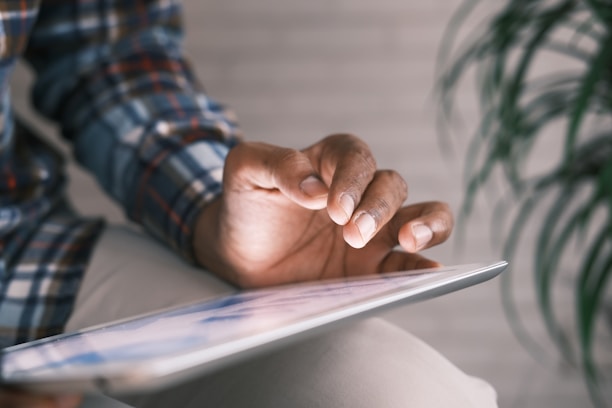 A person engages with a tablet device using their fingertips. The individual is dressed in a plaid shirt, and the focus is on their hand interacting with the touchscreen. A blurred background, featuring an out-of-focus plant, provides a soft context.