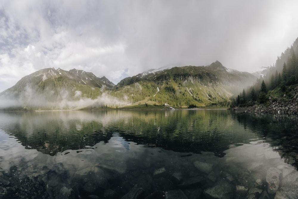 montanhas verdes e marrons ao lado do lago sob nuvens brancas durante o dia