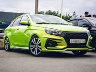 A bright green compact sedan car with sporty features is parked on a street. The car has a sleek design, black accents on the bumper, and stylish alloy wheels. A license plate is visible at the front. In the background, there are trees, a building, and a few people standing nearby.