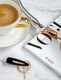 A compact beige crossbody bag placed on a marble countertop with a cup of coffee beside it.