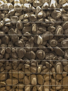 A collection of smooth, rounded stones contained within a grid-like metal mesh structure. The stones vary in size and shape, showing earthy tones and subtle variations in color. The structure appears sturdy, suggesting practical or decorative use.