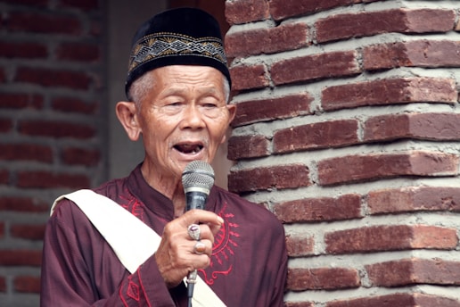 An elderly man wearing a traditional hat and maroon attire holds a microphone while speaking. He stands in front of a brick wall, and his expression is calm and focused.