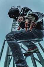A person dressed in dark clothing, including a hood, is crouching on a metal structure. They have mechanical, claw-like gloves extending from their hands, giving a futuristic or cybernetic appearance. The viewpoint is from below, with an emphasis on the person's gloves and legs, set against a cloudy sky.